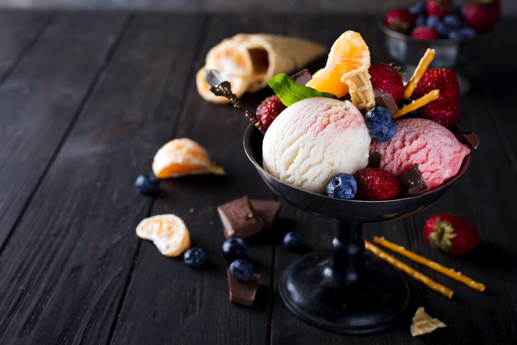 Bowl with ice cream with three different scoops of white, yellow, red colors and waffle cone, chocolate, tangerines and straws