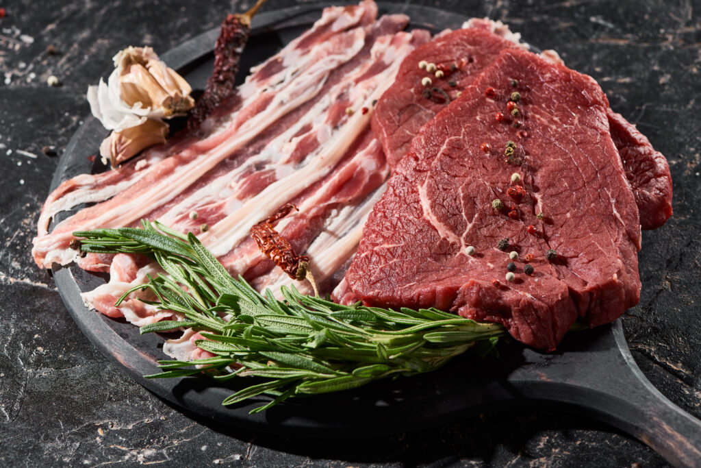 wooden pan with raw beef and bacon near rosemary, garlic and cayenne pepper on black marble surface